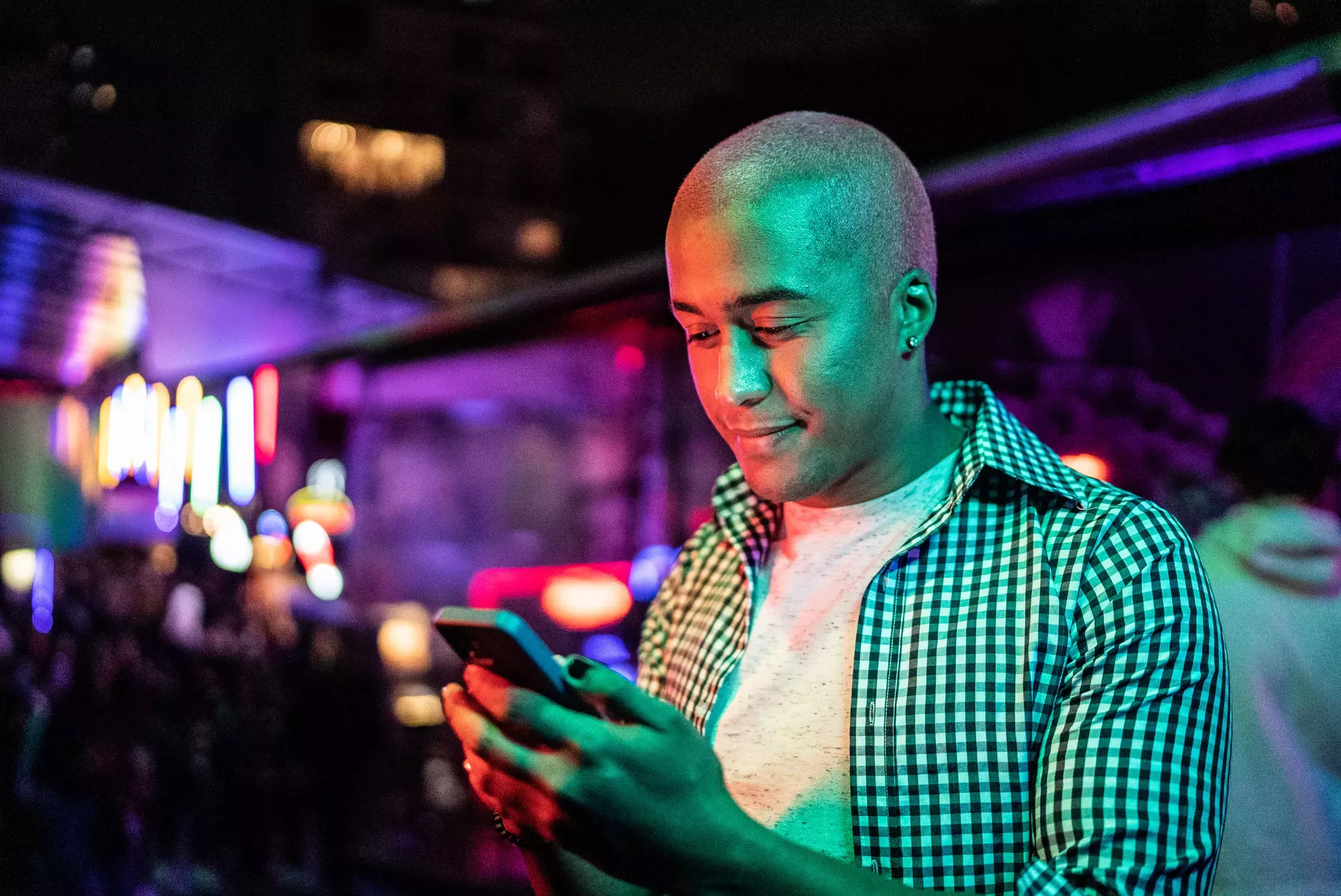 A young man with a buzzcut wears a white crewneck t-shirt under a plaid button down shirt unbuttoned. He stands looking at his iPhone, which he holds in his left hand. He has pierced ears and painted black nails. He stands in the midst of a purple-colored gay bar, blurred in the background.