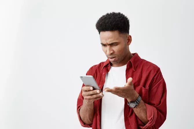 Close up portrait of young good-looking man in casual white t-shirt with red shirt looking at phone with confused expression