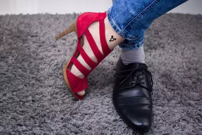 Person has a lady's boot on one foot, and on the other a male boot.The person has a third-gender tattoo.