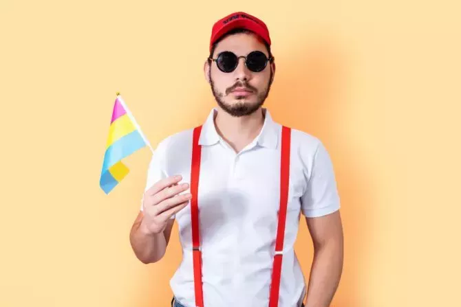 Young hipster holding Pansexual pride community flag, on light orange background.