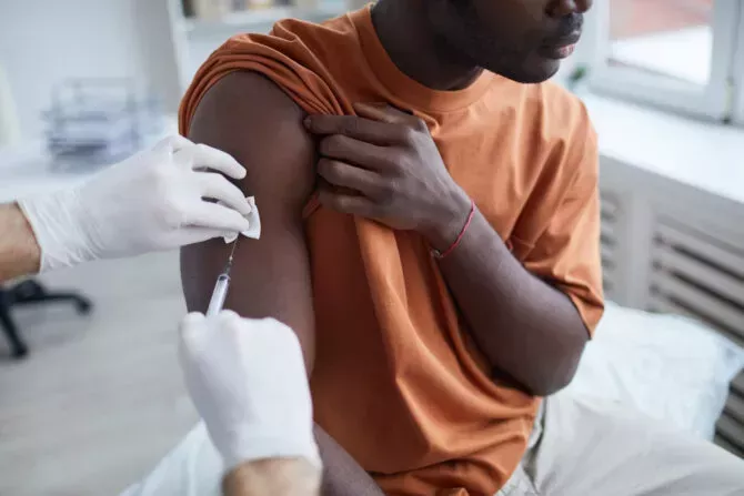 Close-up of adult African American man looking away while receiving covid vaccine at clinic or hospital, with male nurse injecting vaccine into shoulder