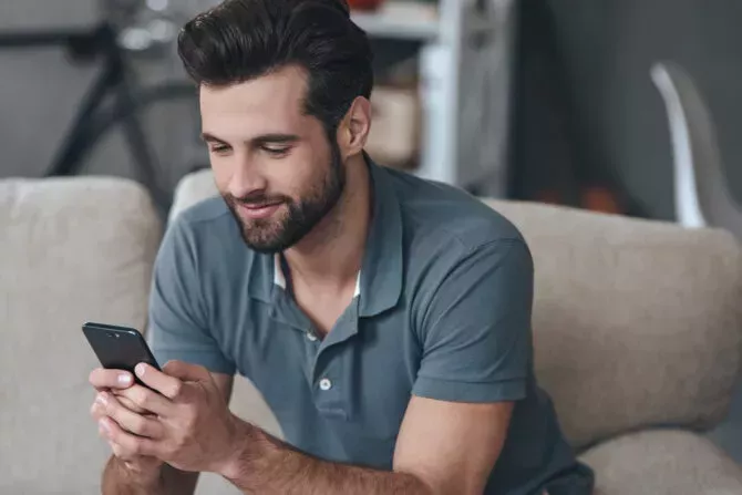 Cheerful handsome young man using his smart phone while sitting on the couch at home