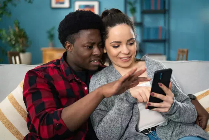 young couple sitting on the grey couch in the cozy room while watching something on the cell phone device and talking at home