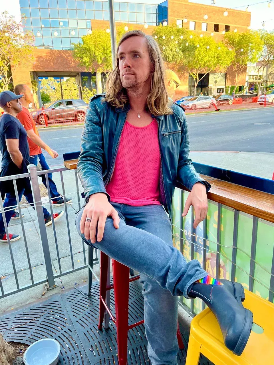 Genderfluid individual sitting outside a bar in Los Angles, their legs are crossed and they are wearing rainbow boots