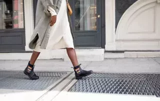 Blundstones Pride boot Lower half of a pedestrian on a city street, walking across the pavement. They are in a white bubble dress with chelsea style boots with rainbows inlaid on the sides.