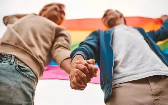 Two men hold hands in front of a rainbow flag