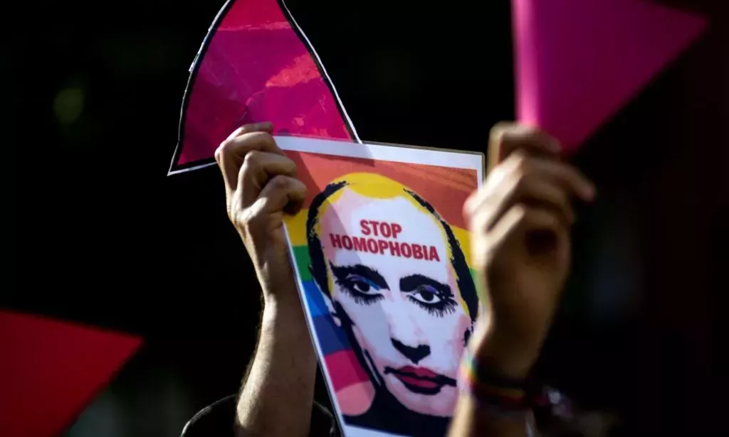 A person holds up a sign with a depiction of Russian president Vladimir Putin with LGBTQ+ rainbow colours in the background with the words 'Stop homophobia' across his forehead
