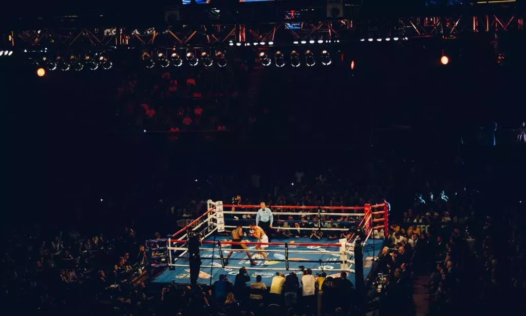 A photo taken from where people sit in the arena shows two people fighting against each other in a boxing ring