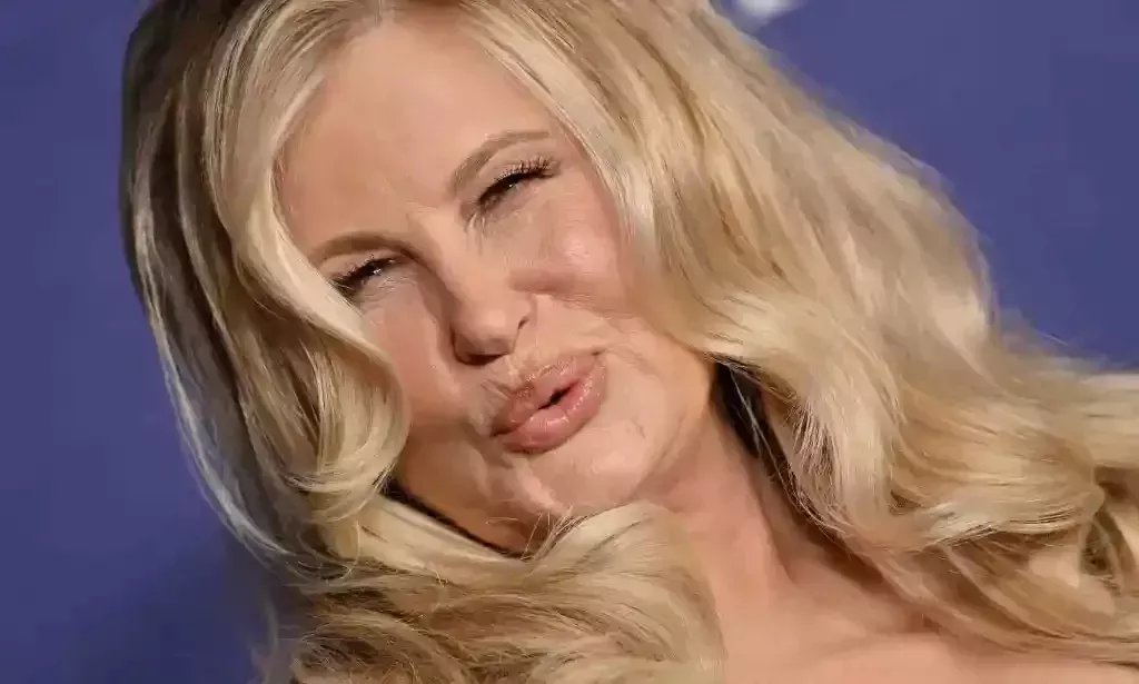 A cropped image of American actor Jennifer Coolidge with long blonde hair standing in front of a blue background and pouting