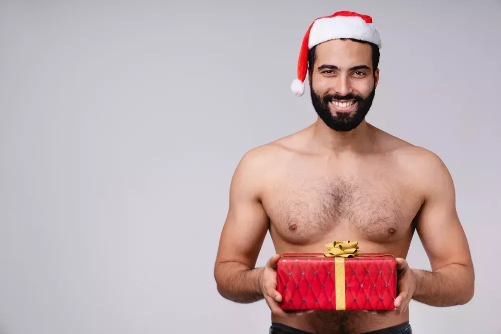Shirtless man in a red Santa hat holding a Christmas present.