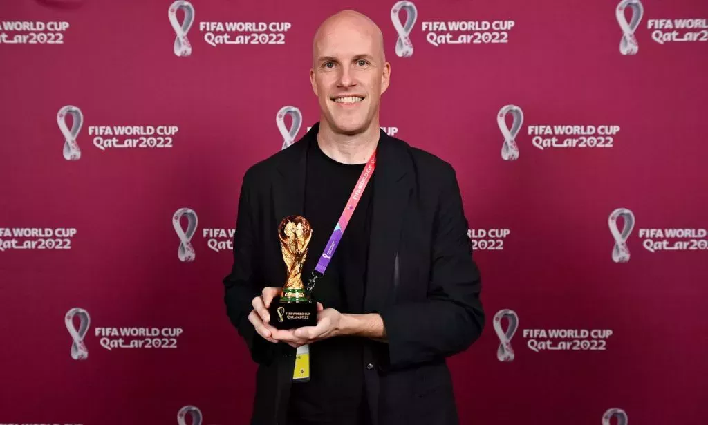 Grant Wahl stands infront of a red back drop with the Qatar World Cup logo written across it, while holding a miniature replica of the World Cup trophy, wearing a black blazer.