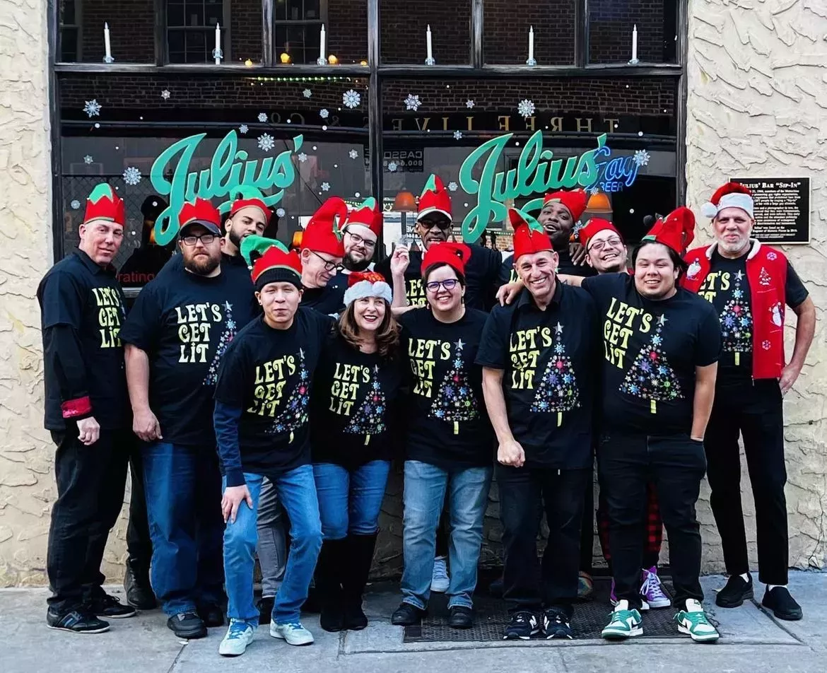 A crowd of folks in Christmas shirts and elf hats stand in front of Julius' bar in NYC.