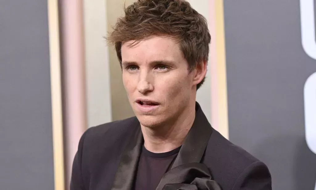 Eddie Redmayne wears a brown shirt and matching brown jacket as he poses for the camera