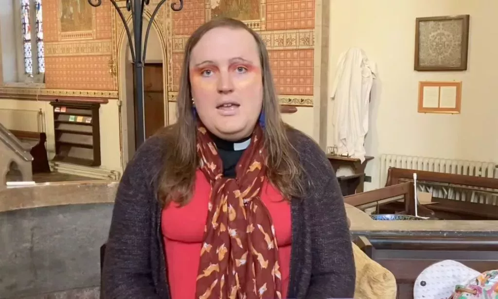 Bingo Allison, a non-binary, genderqueer Church of England priest, wears a red top, sweater and scarf while they speak about religion and Trans Day of Visibility