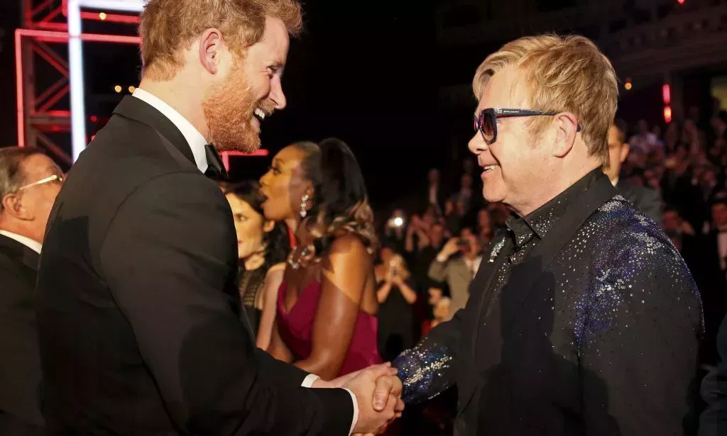 Prince Harry, wearing a suit and tie, shakes the hand of Elton John, who is wearing a dark suit with a black shirt, at a public event
