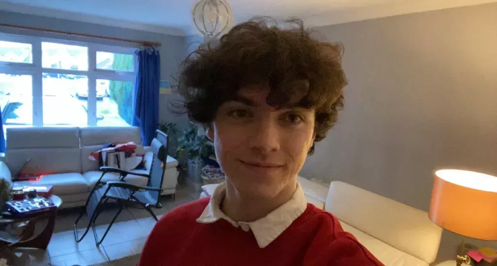 Josh McGiff, a 22-year-old gay man, is pictured in a selfie with a red shirt and dark hair in an internal room with a sofa in the background.