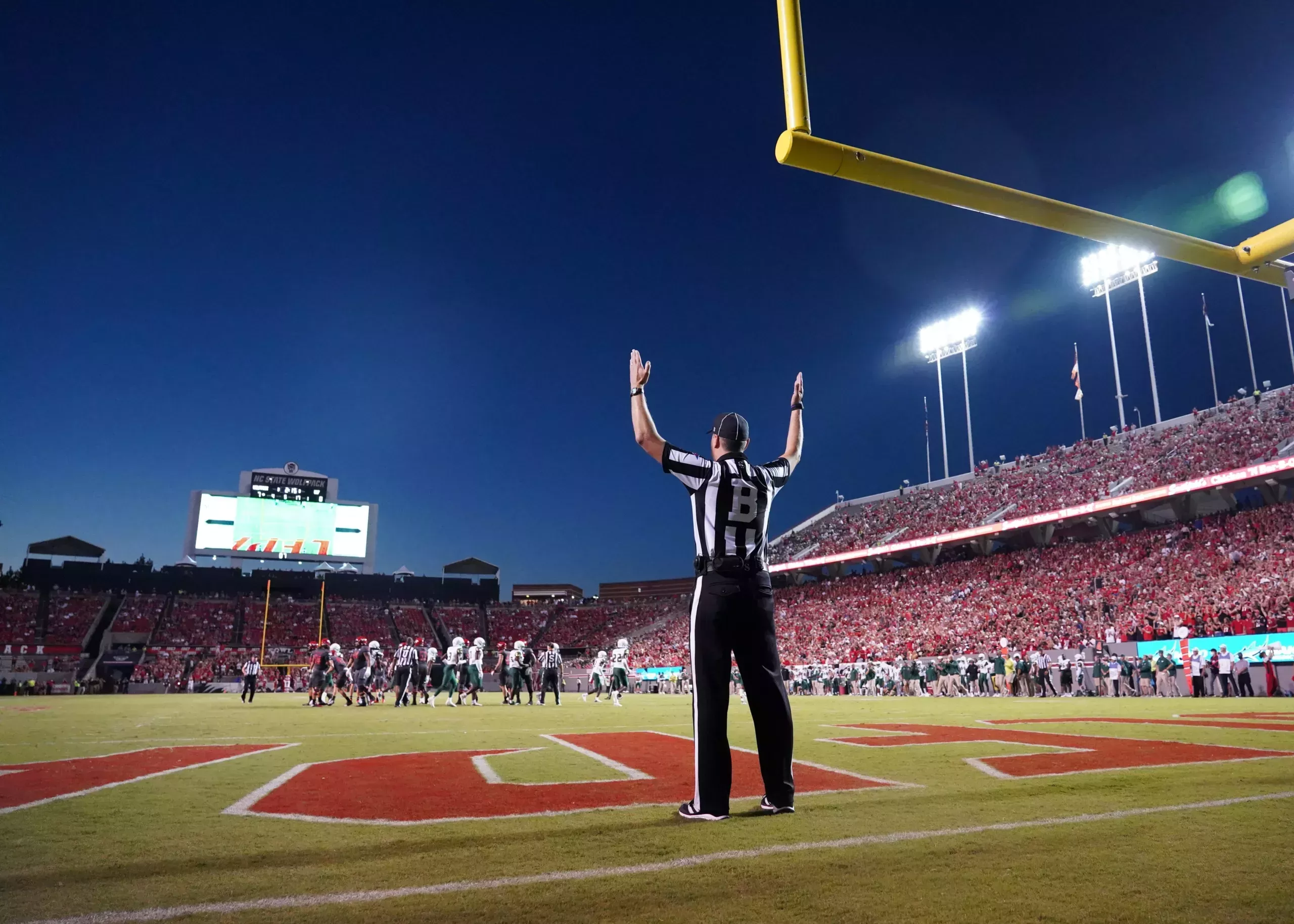 A referee standing in an end zone under a field goal post with their arms raised
