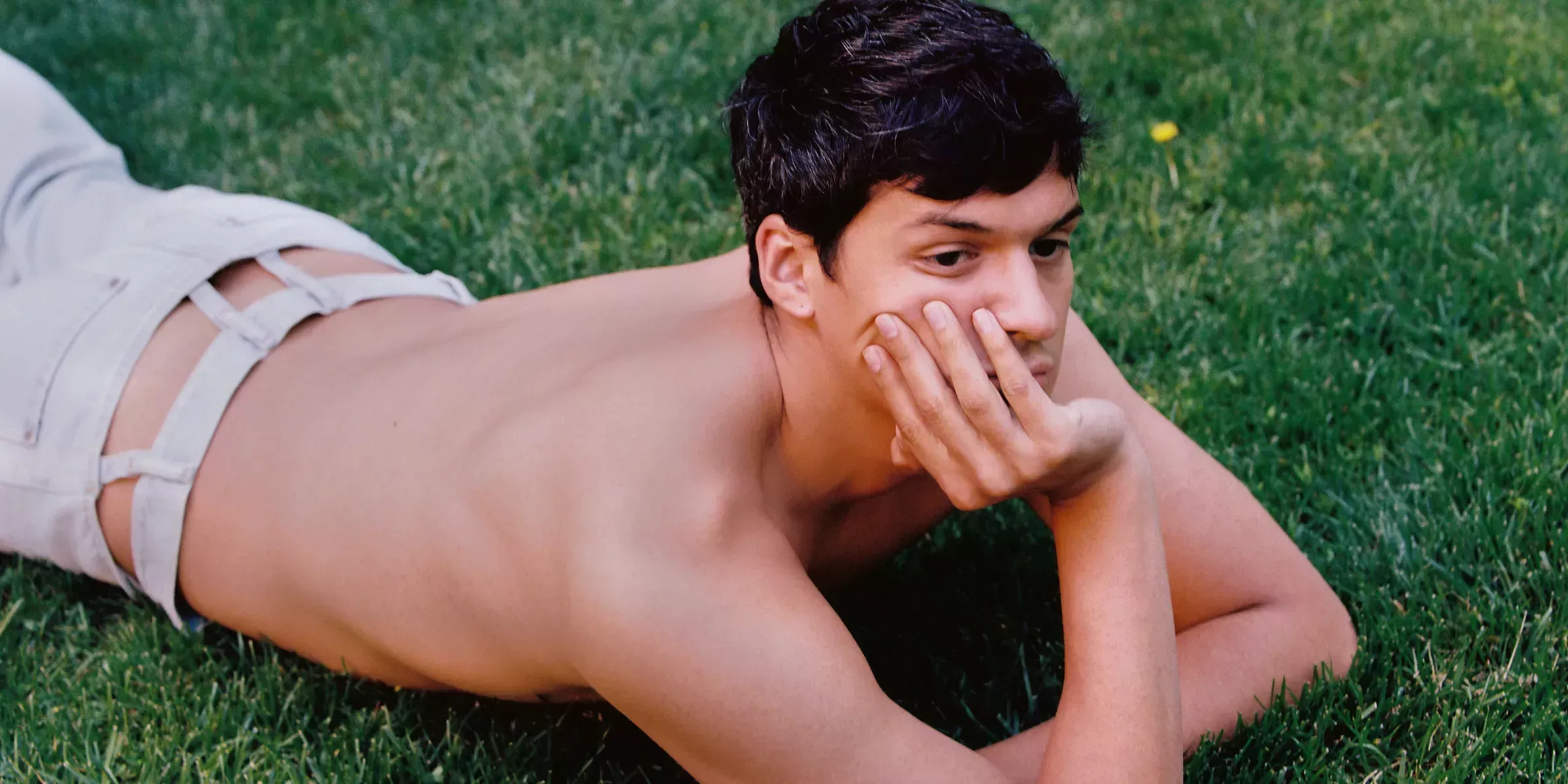 Omar Apollo lying shirtless in a field of grass.