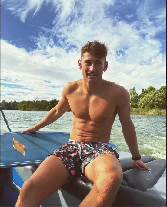 Jake Williamson pictured wearing swimming trunks on a boat. In the background the water is visible and the sky is blue with clouds swirling.