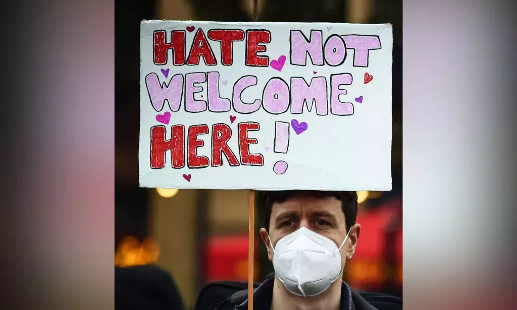 A person wearing a white face mask holds up a sign reading "hate not welcome here!" during a protest in support of the LGBTQ+ community and trans people