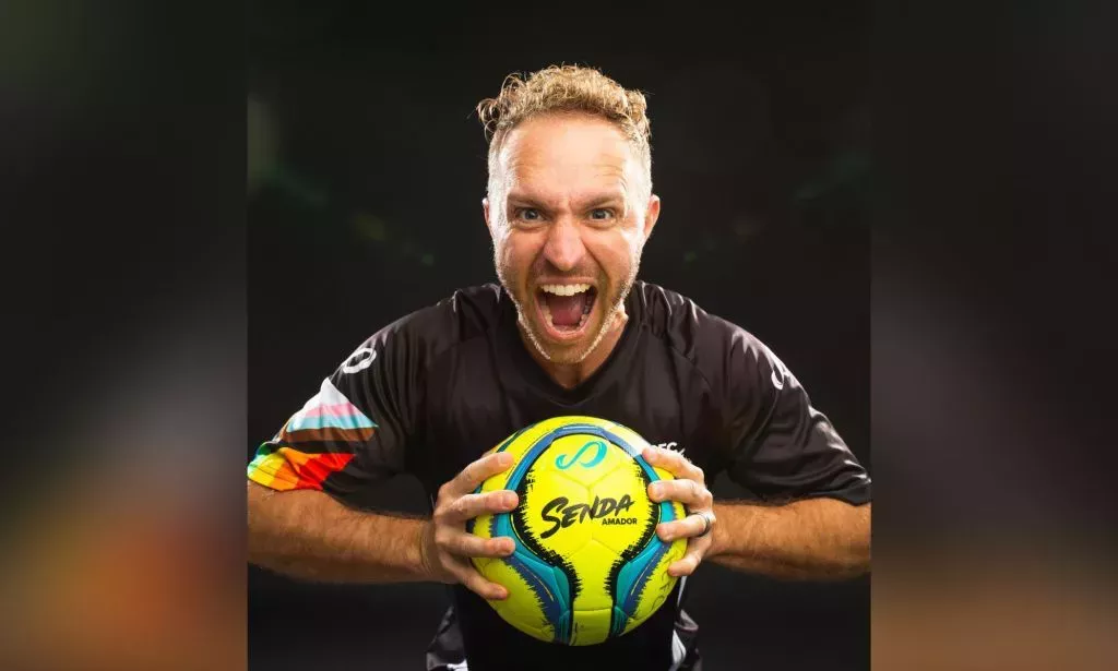 Openly trans coach Kaig Lightner wears a black football jersey with a progressive pride flag on one shoulder as he holds a football in his hands
