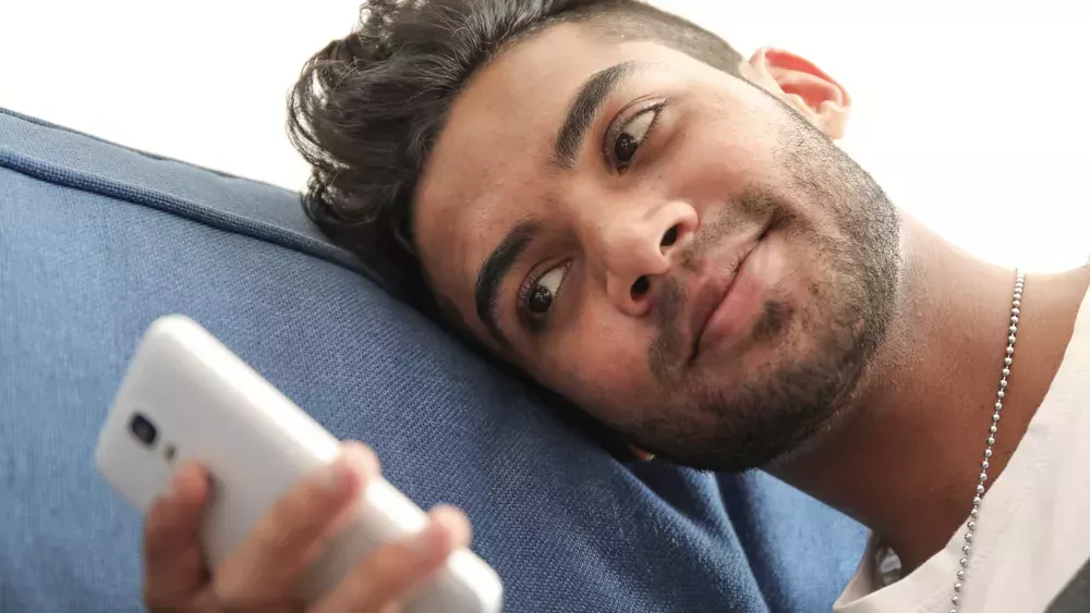 Bearded man lays on his bed and looks at his cellphone.