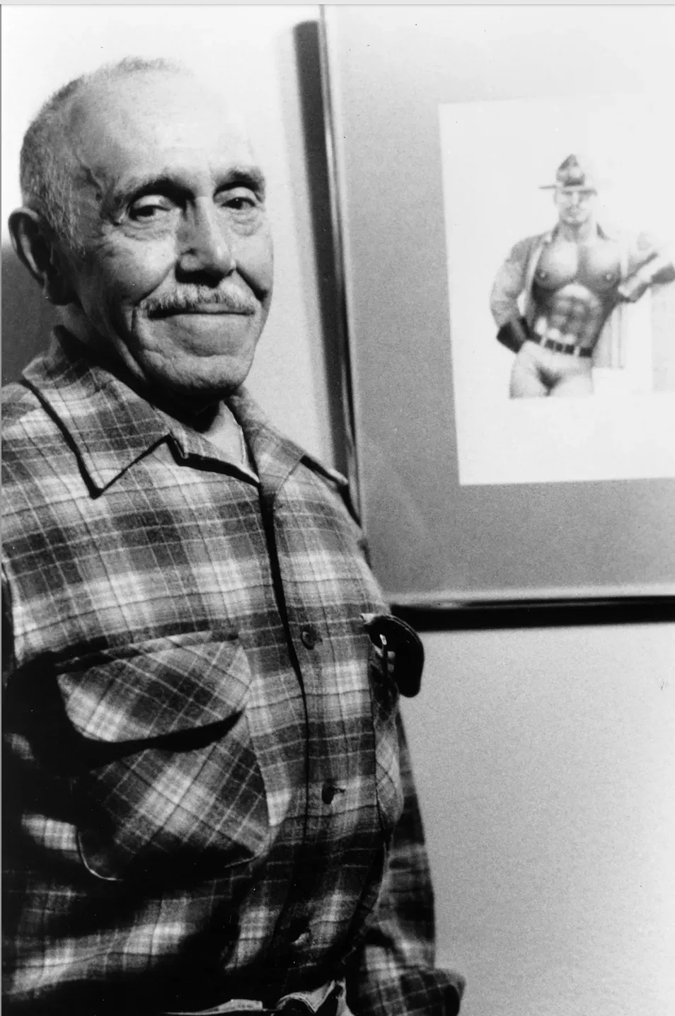Tom of Finland smirking, standing in front of one of his drawings. 