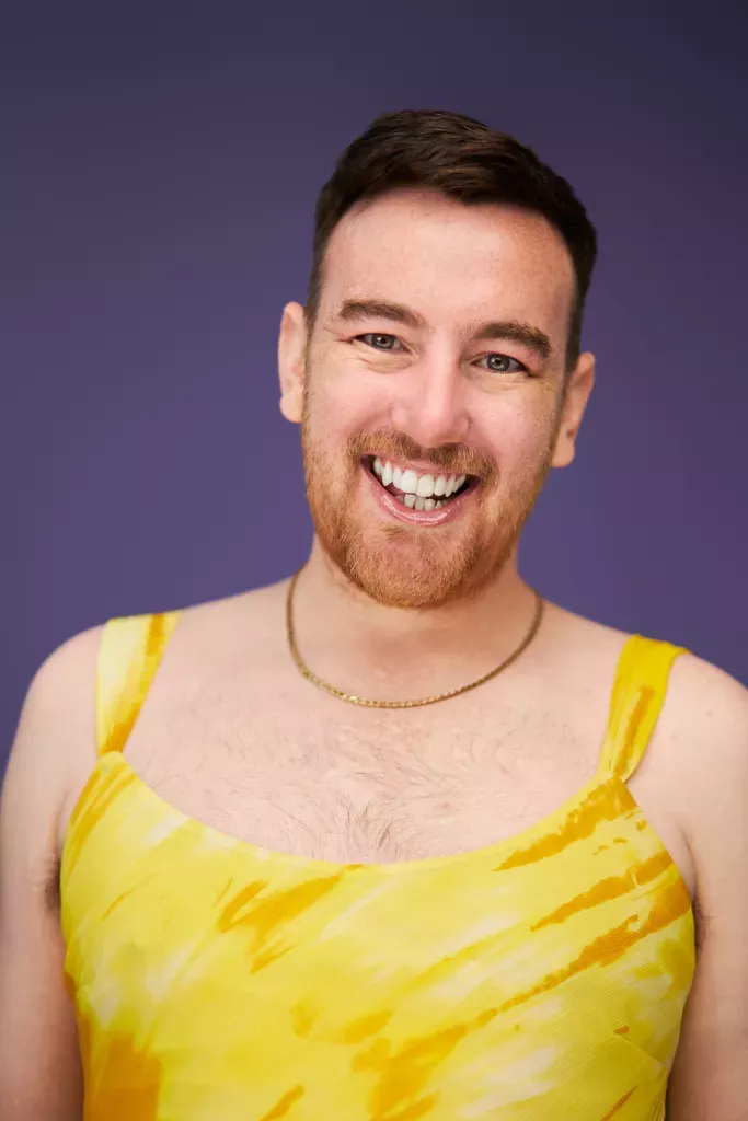This is an image of Chris King. They are wearing a brigh yellow dress. They have a reddish coloured beard and are smiling. 