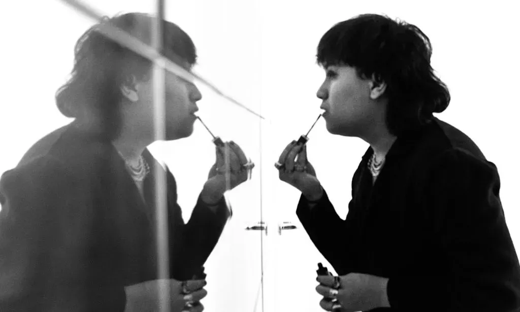 A black and white photo of non-binary, Malaysian singer Shaf (aka moreofthem) wearing dark clothes as they look at themself in the mirror and put makeup on their lips