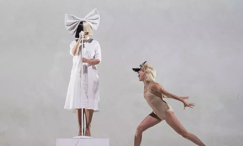 Sia performs in a white dress and blonde and black wig, which covers her face. Maddie Ziegler dances next to her.