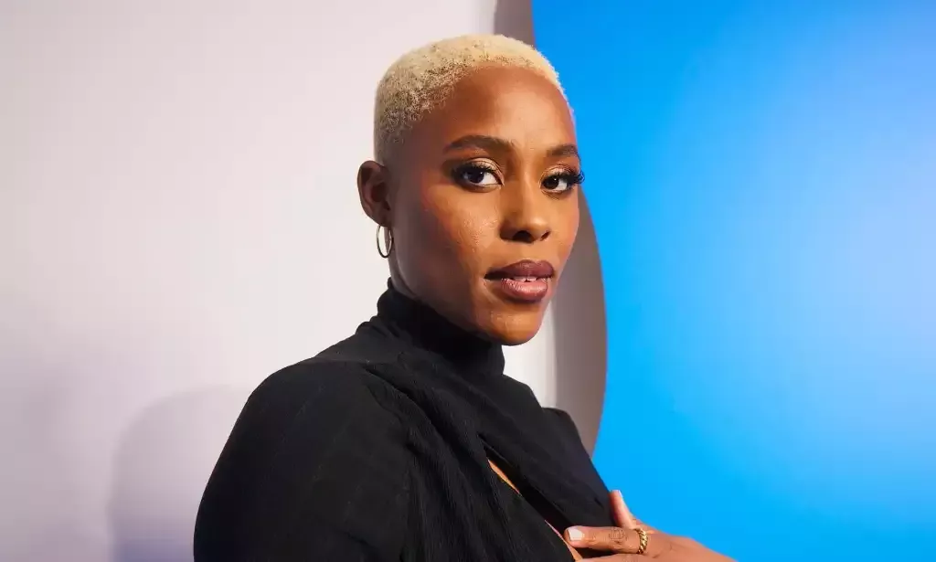 A promo shot of actor Jerrie Johnson wearing a black dress with short white-blonde hair standing in front of a blue and white background