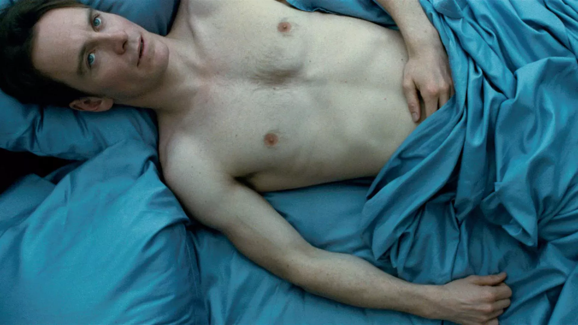 Michael Fassbender laying in bed shirtless. He is covered by blue sheets. 