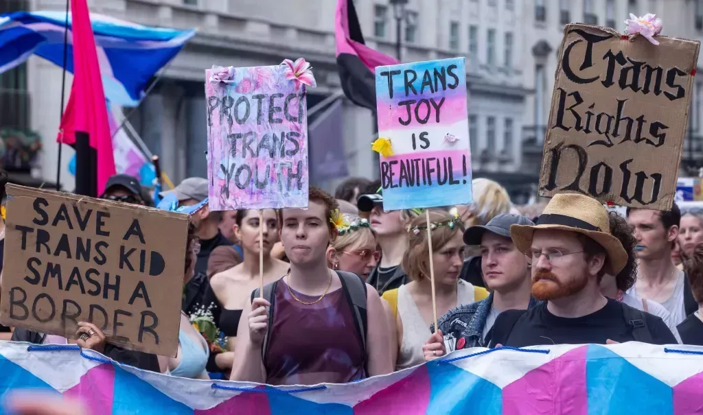 People walk down the street during a Pride march with signs reading "trans joy is beautiful" and "protect trans youth"
