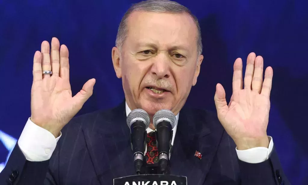 Turkey's president Recep Tayyip Erdoğan, who has a history of making anti-LGBTQ+ remarks, wears a suit and tie as he holds up both his hands while speaking into a microphone