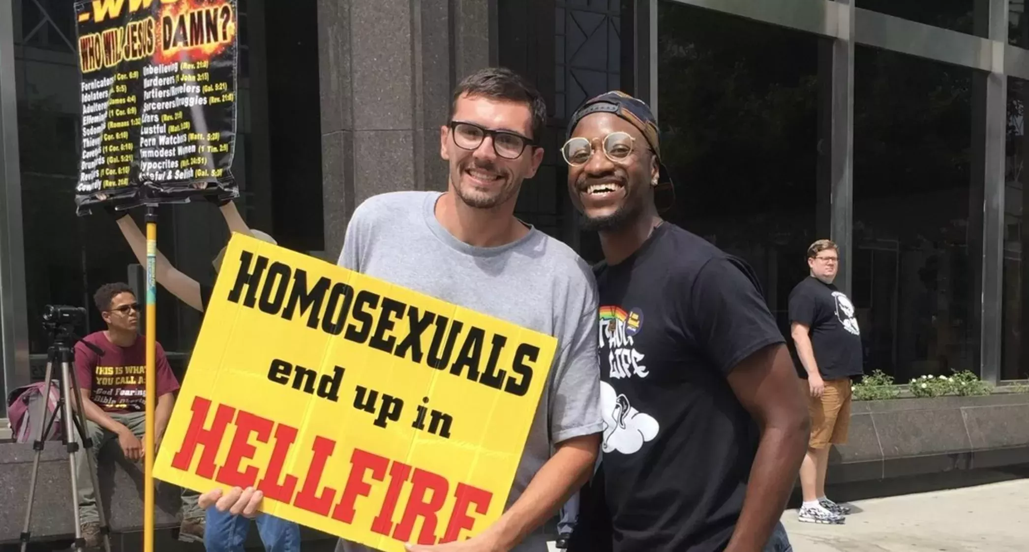 A white man in a gray shirt and glasses holds up a yellow sign that reads "Homosexuals end up in hellfire" while smiling next to a gay Black man in a backwards baseball hat, glasses, and a black shirt with a rainbow on it. They stand smiling outside in front of a windowed building while onlookers watch.