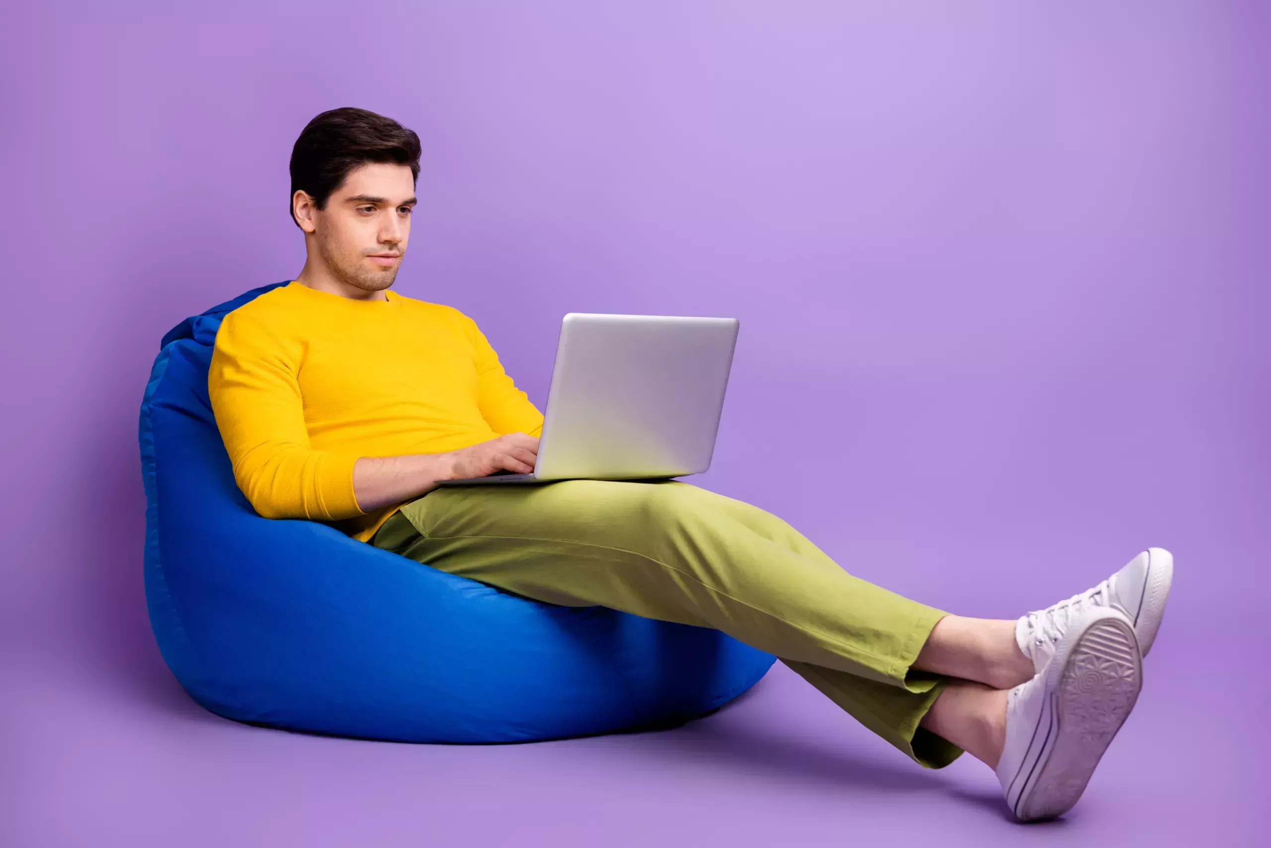 A man wearing a yellow shirt on the computer and sitting on a bean bag. 