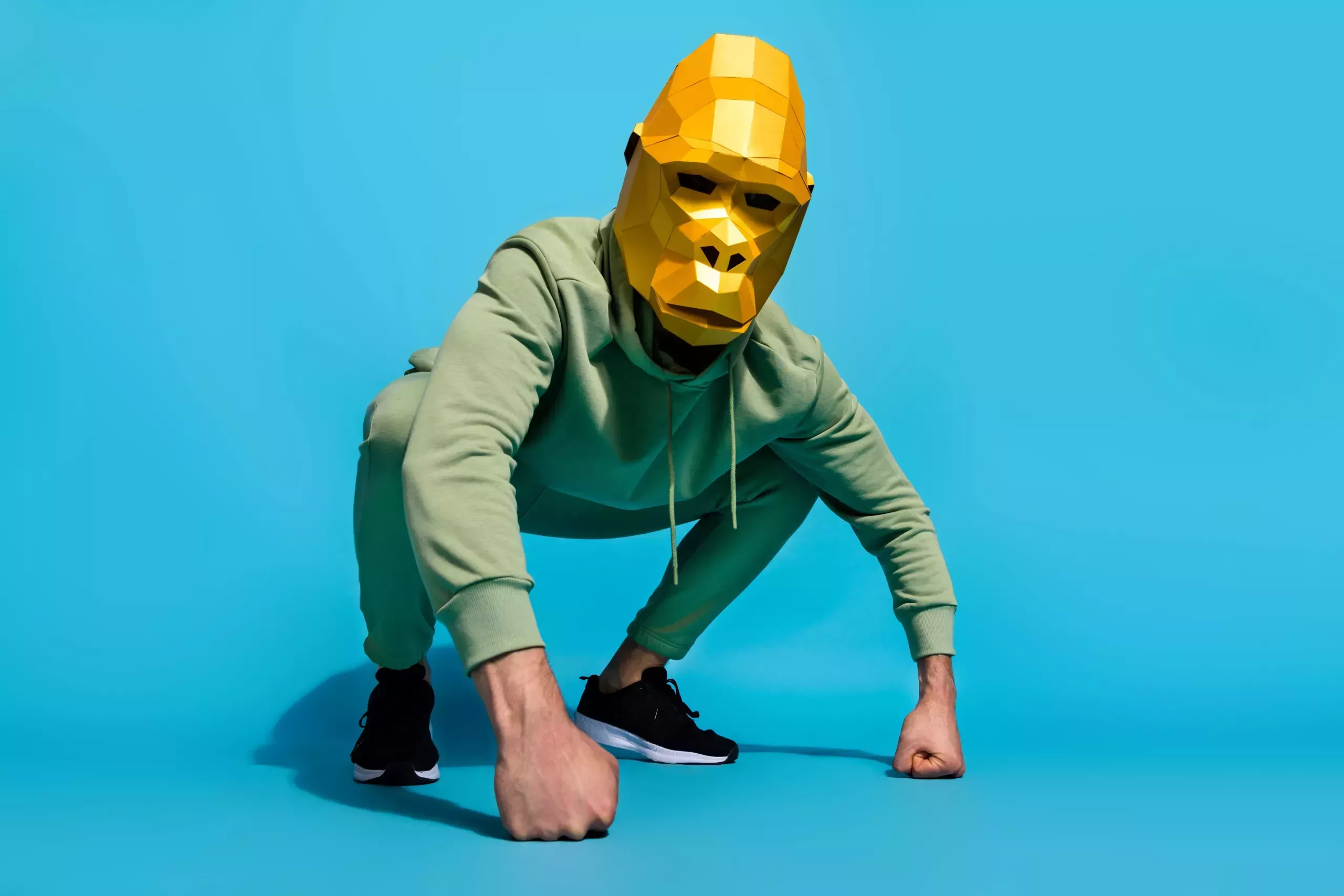 A mean wearing green sweats and a golden gorilla mask kneeling down with his fist on the floor. 