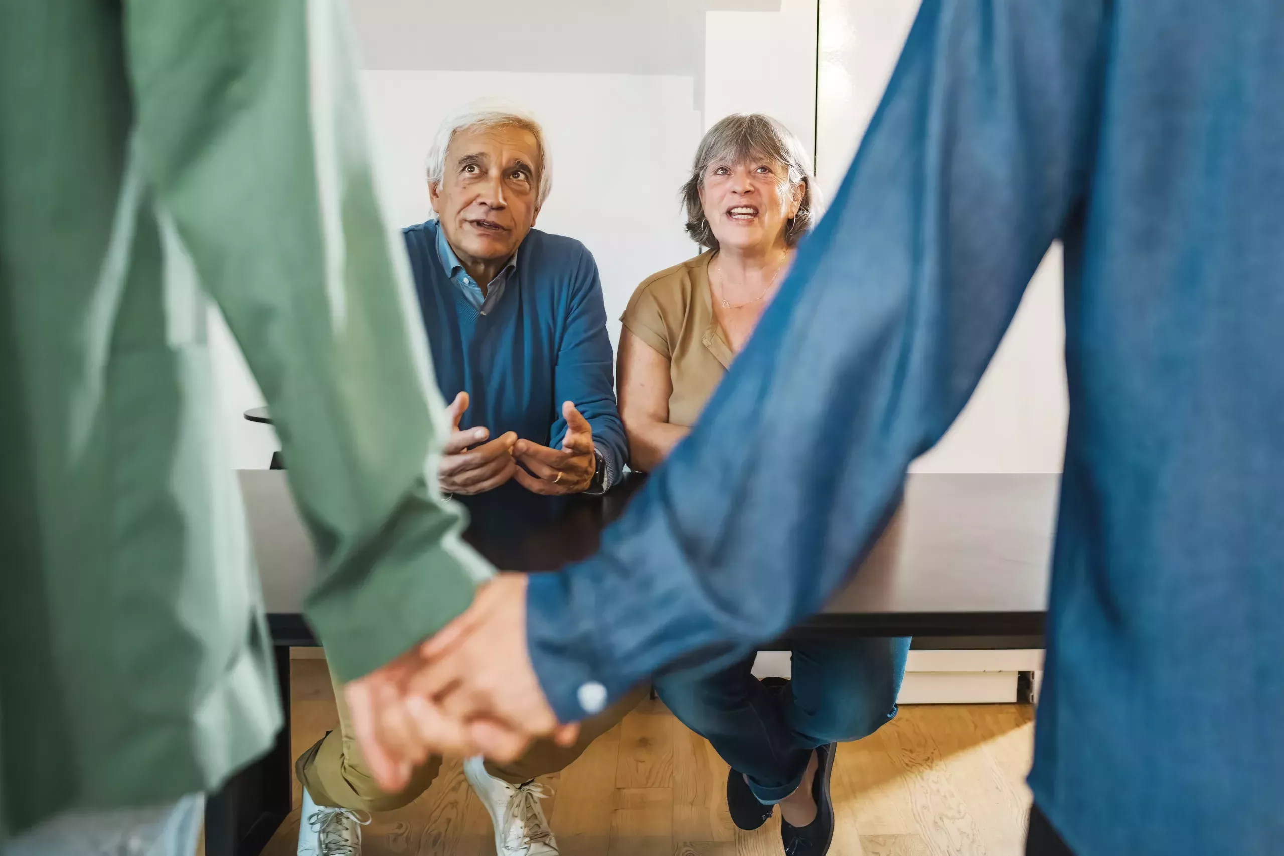 A man in a long sleeved green shirt holds hands with a man in a long sleeved blue shirt. Their backs face the camera, and in the background, an older man and woman sit at a black table looking at them quizzically.
