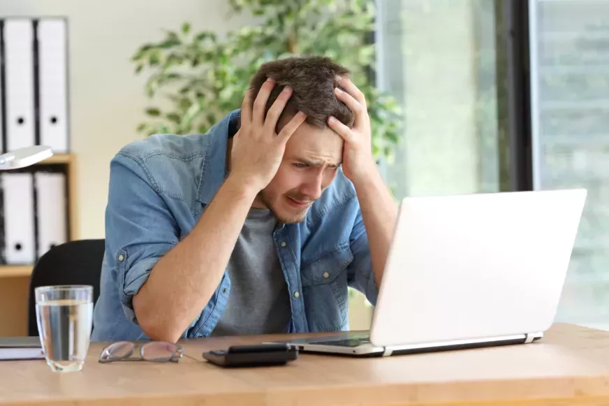 Man in denim shirt looking devastated at table while looking at a laptop.