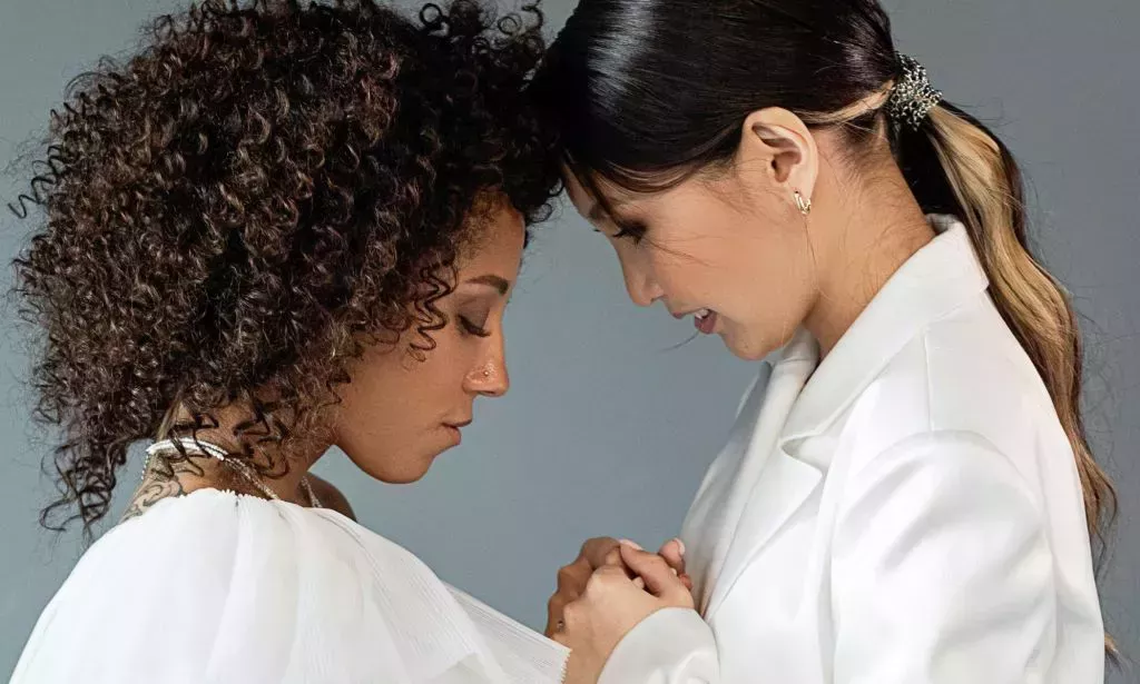 A queer, same-sex couple wear white outfits as they attend a wedding and hold each other's hands