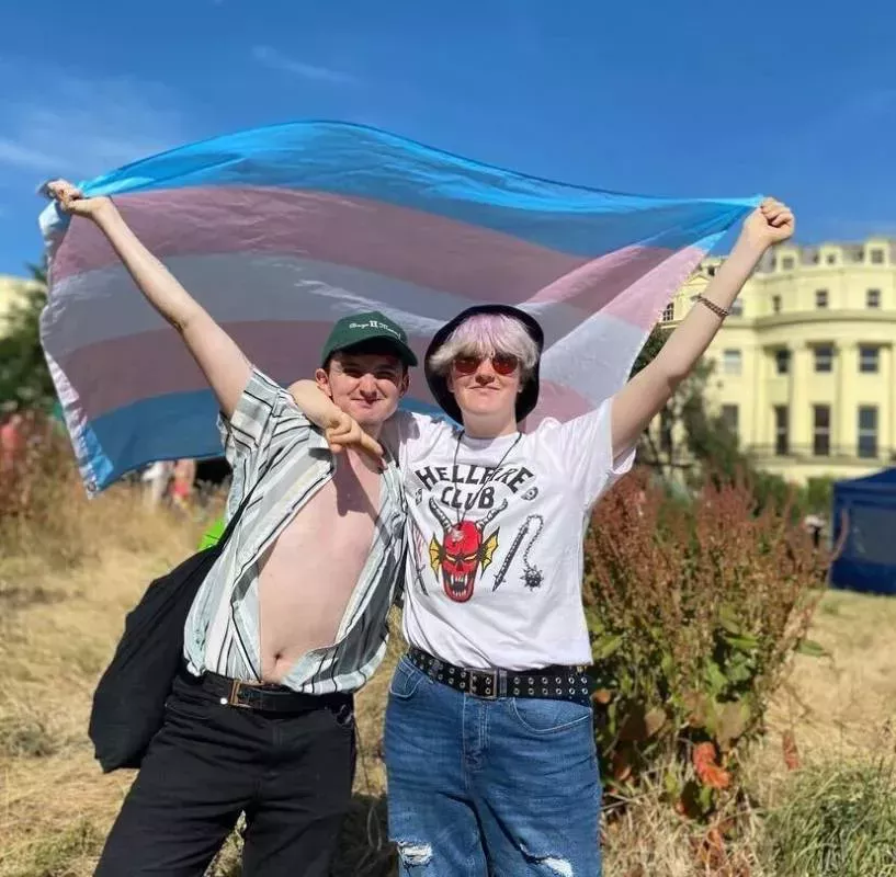 Oli, who has top surgery, and his non-binary sibling hold up a trans Pride flag while standing on a Brighton beach