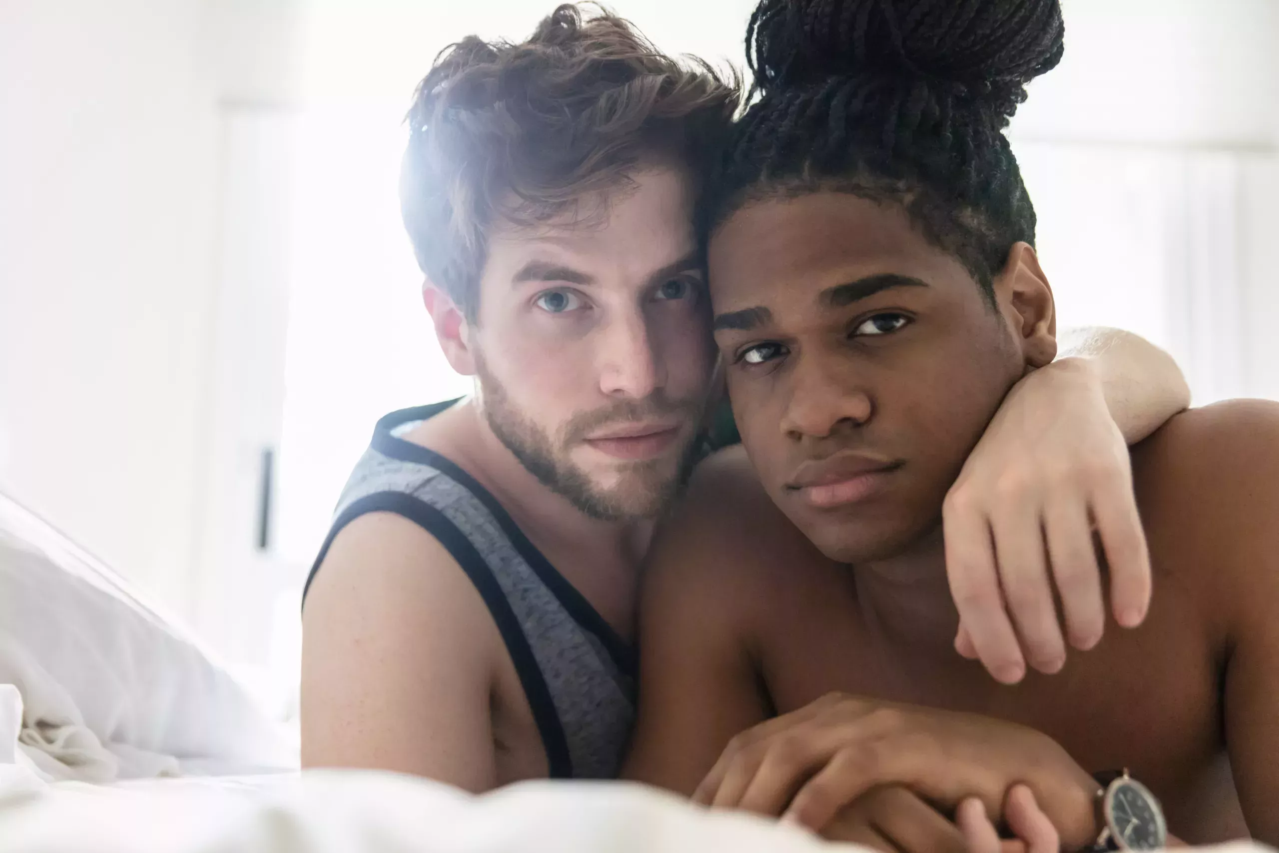 Diverse homosexual couple lying down on bed looking at camera very serious