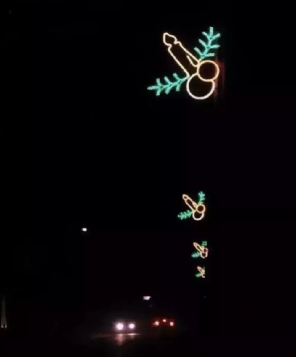 A dark street lined with neon Christmas decorations. Each neon decoration is a candle in between two ornaments and a sprig of holly, unfortunately resembling a penis. 