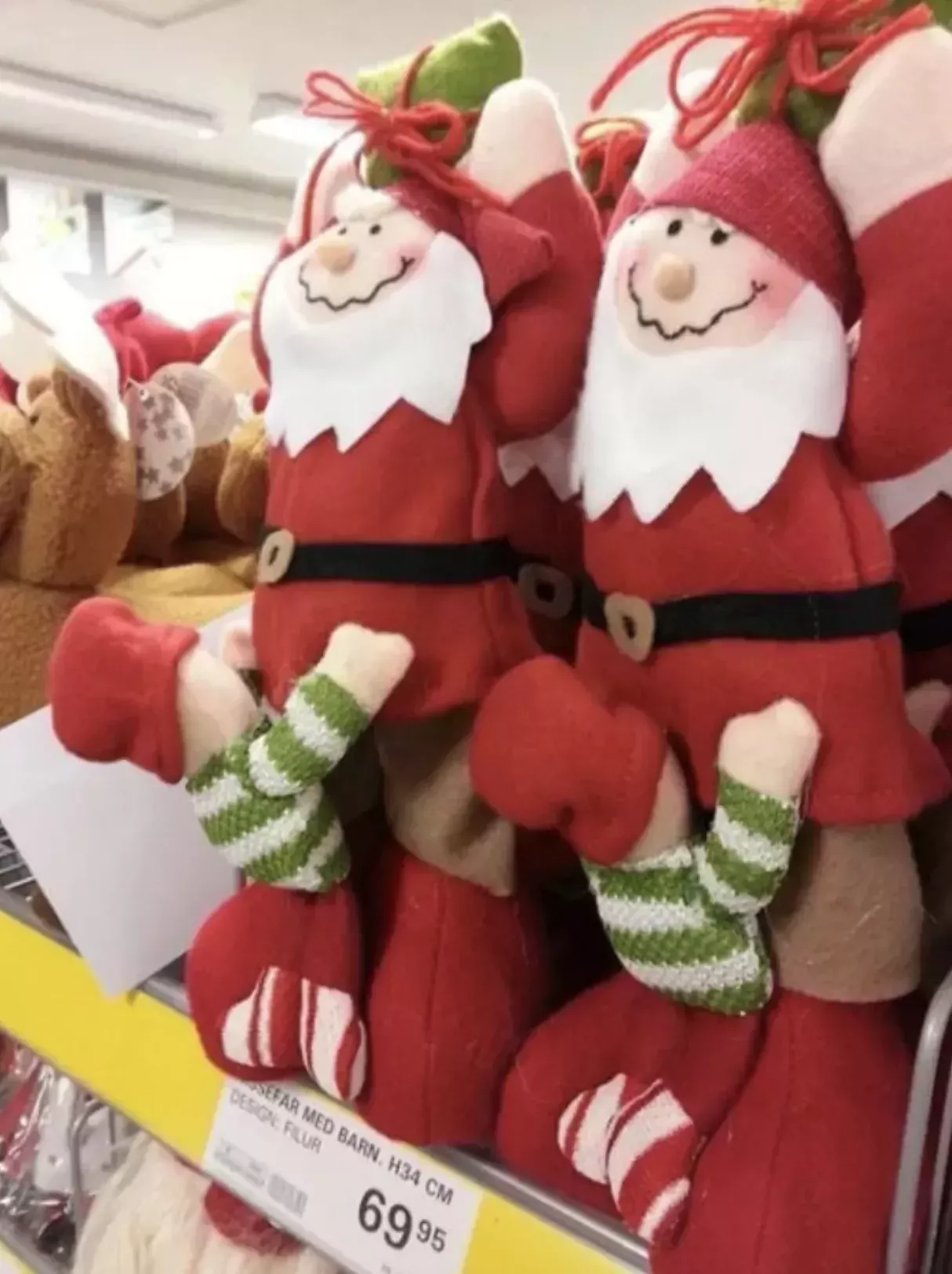 A collection of stuffed Santa decorations on the shelf at a store. The decoration features Santa with a smile holding a present above his head. Sewed to the front of his crotch, unfortunately placed between his legs, is a little child with his face buried in his crotch. The child wears a green striped shirt and candy cane striped shoes.