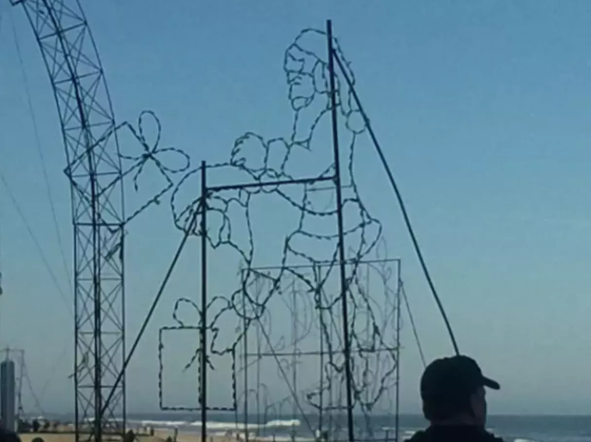 On the beach, a Christmas light decoration pictured at daytime. What is supposed to be a Santa that lights up and gives the appearance of a tossing up a present now looks like one Santa thrusting into another who's bent over.