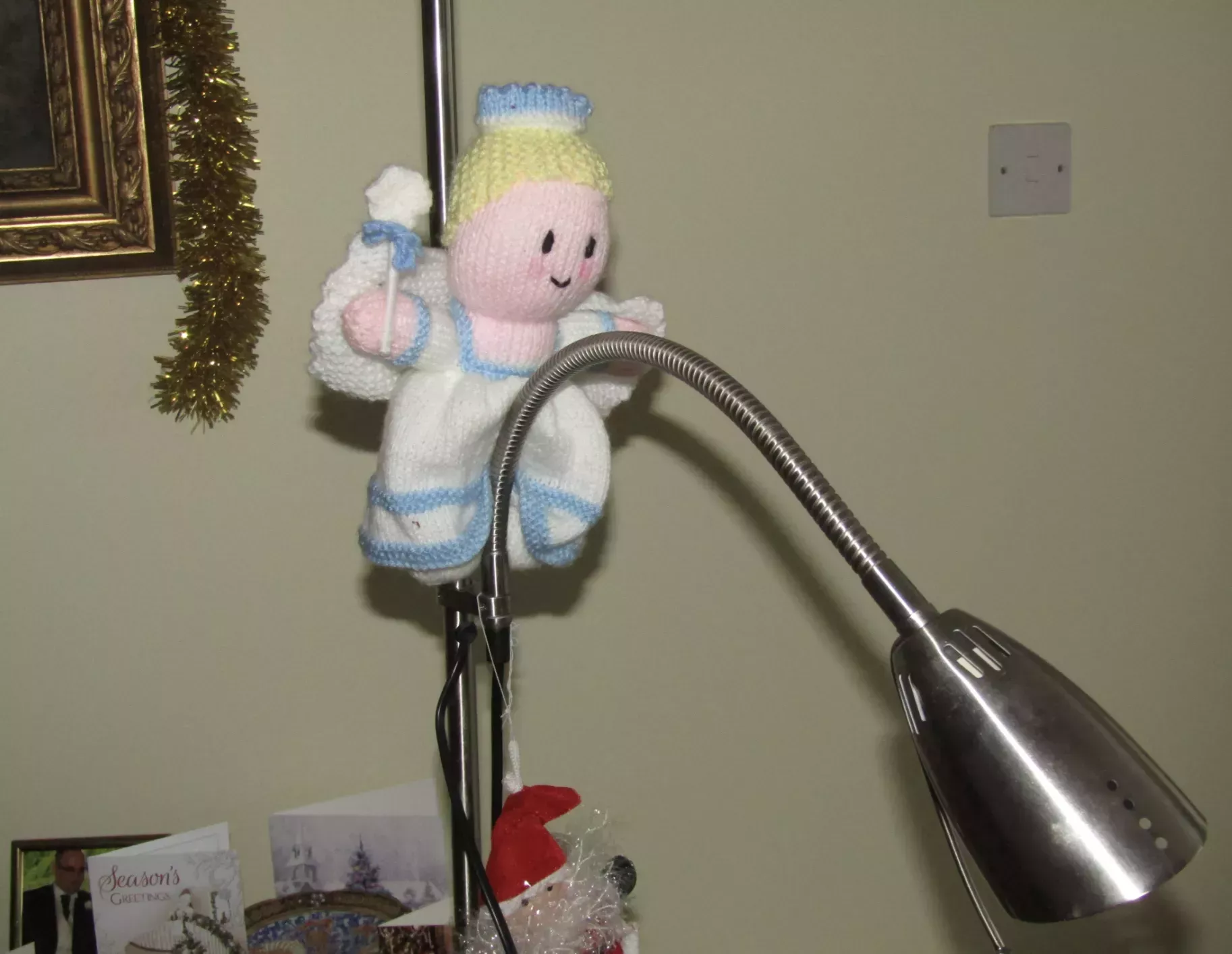 A crocheted blonde angel in a white gown with white wings and a lollipop is tied up on a desk lamp, which hangs between its legs resembling a phallus. In the background are Christmas cards and holly decorations.