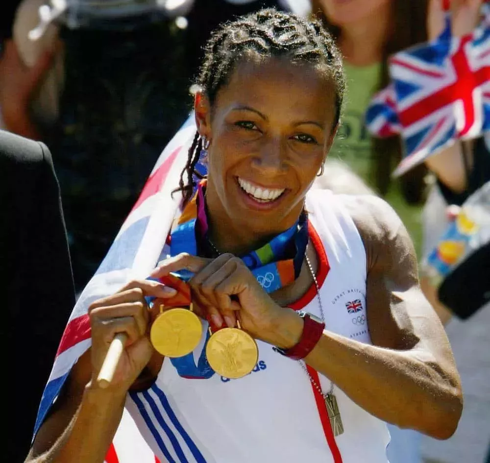 Dame Kelly Holmes wears a white red and blue Olympic outfit as she holds up two Olympic gold medals and clutches a Union Jack flag in her hand
