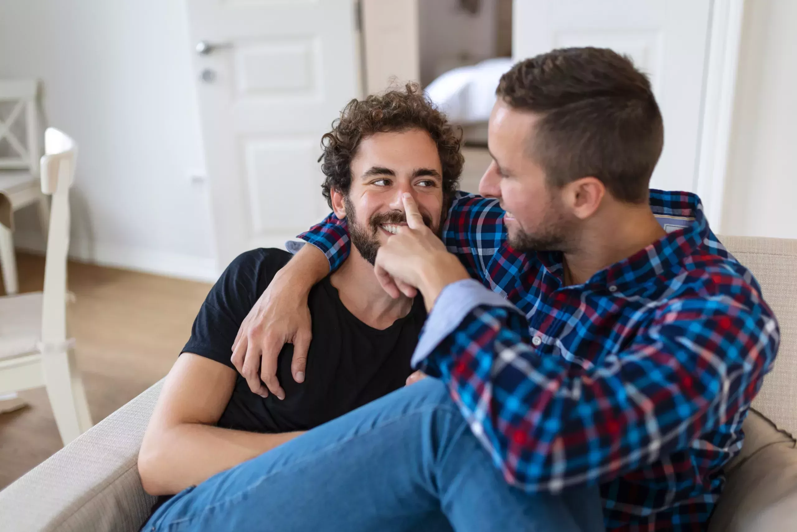 Two men sharing a close and joyful moment on a couch, with one man playfully touching the other's nose. They are both smiling and seem to be engaged in a lighthearted and affectionate conversation, creating an atmosphere of comfort and intimacy. Their queer love language would be physical touch.