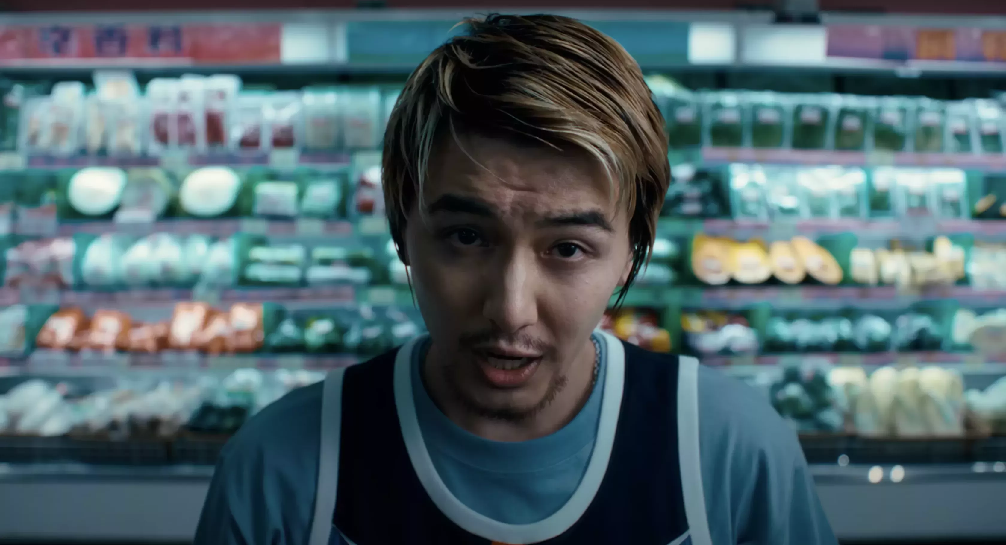 J-pop singer Fujii Kaze, with long bleached hair and a thin mustache smiles while performing in the music video for "Workin' Hard." He wears a navy jersey over a light blue t-shirt and stands in the middle of a grocery store, with the produce aisle blurred behind him.