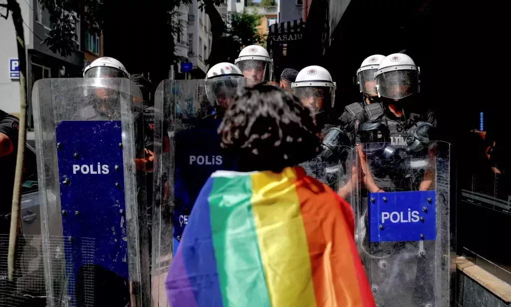 Una persona LGBTQ+ con una bandera del Orgullo sobre los hombros enfrent&aacute;ndose a la polic&iacute;a en Turqu&iacute;a.